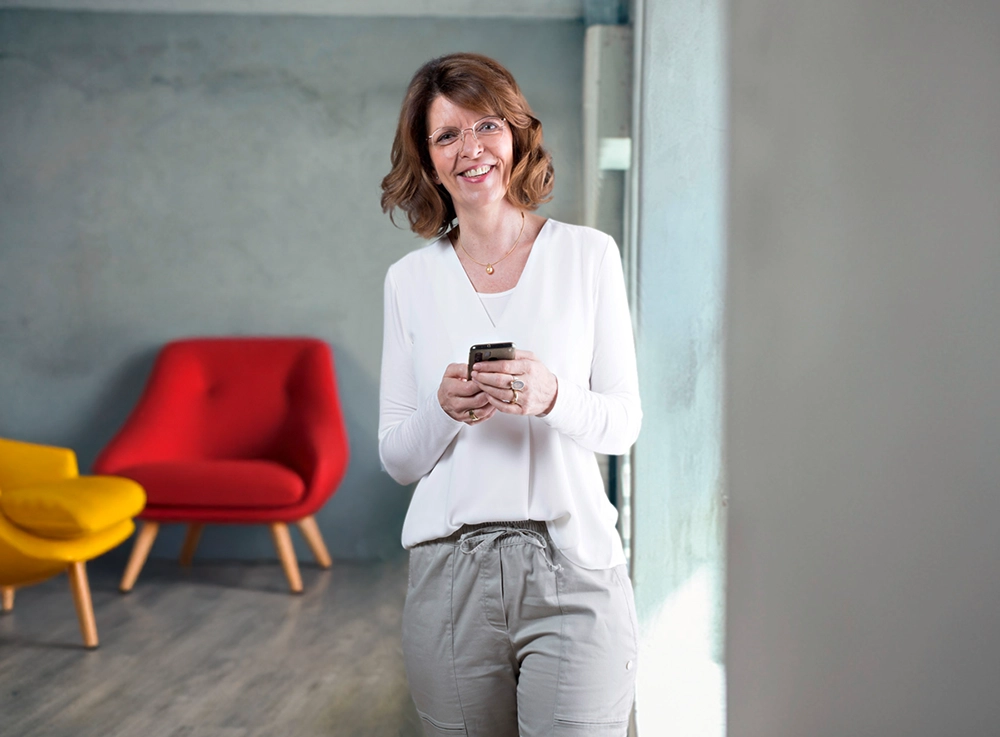 Gabriele Aumeier steht entspannt mit Smartphone in der Hand, im Hintergrund bunte Sessel.