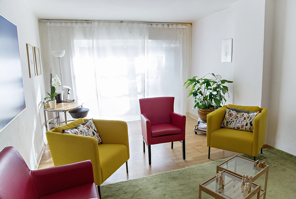 Ein heller Raum mit einer gemütlichen Sitzgruppe aus gelben und roten Sesseln, um einen Glastisch arrangiert, dekoriert mit Pflanzen und Kunstwerken.
